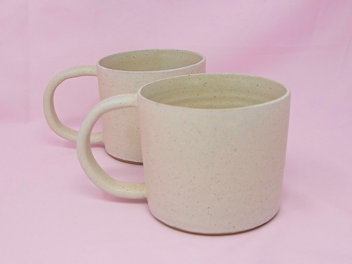A pair of handmade cream speckled mugs against a pink background.